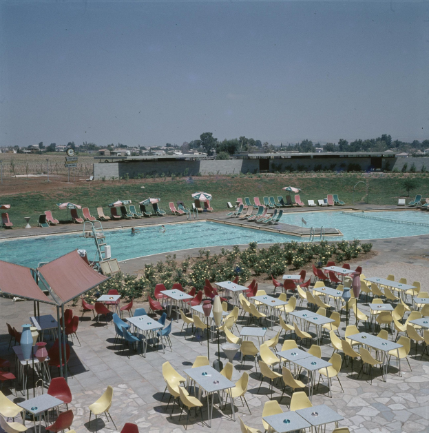 Ein Freibad mit Tischen, Stühlen und Sonnenschirmen, umgeben von Blumenpflanzen, einem Zaun, einer Wand, Bäumen, Gebäuden und einem bewölkten Himmel.