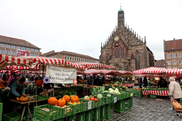 Ein belebter Markt in Nürnberg, Deutschland, mit verschiedenen Obst- und Gemüsesorten, Menschen mit Taschen und um den Markt aufgestellte Zelte, mit Gebäuden und einem Uhrenturm im Hintergrund unter einem sichtbaren Himmel.