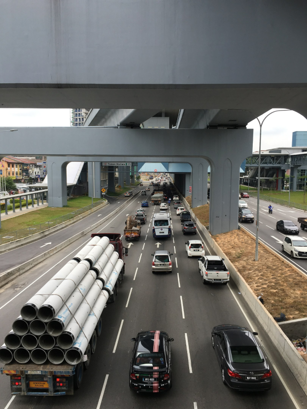 Eine vielbefahrene Autobahn mit mehreren Fahrzeugen, Verkehrszeichen, Straßenlaternen, Gras, Gebäuden und Bäumen unter einer Brücke, mit dem Himmel darüber.