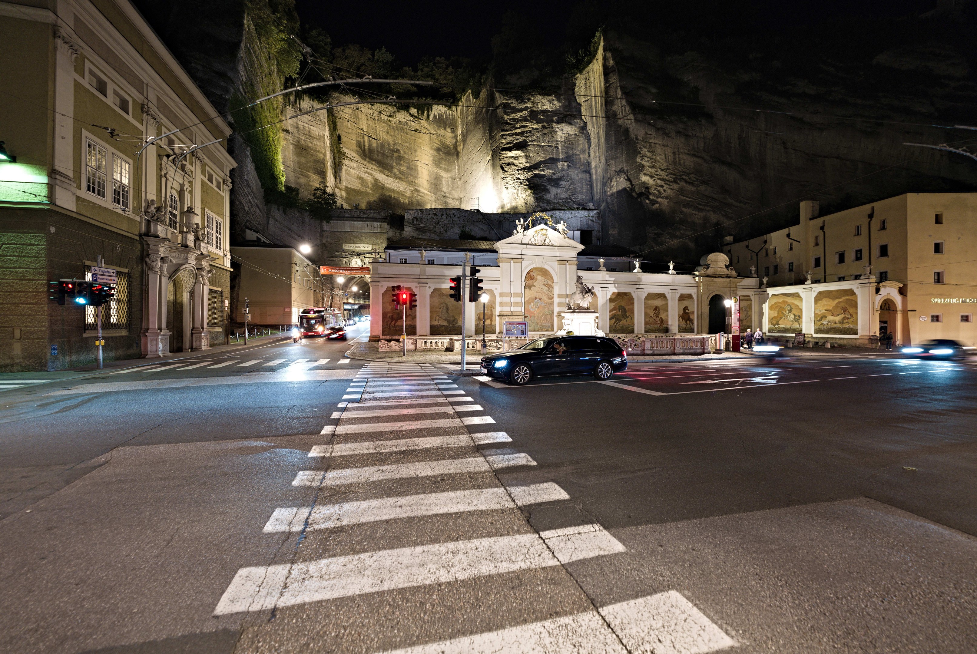 Eine nächtliche Straßenansicht mit fahrenden Autos, Straßenlaternen, Verkehrsampeln, beleuchteten Gebäuden, Laternenpfählen, einem Hügel im Hintergrund und einer Zebrastreifenmarkierung unten.