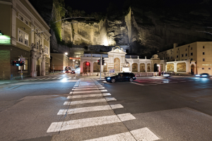 Eine nächtliche Straßenansicht mit fahrenden Autos, Straßenlaternen, Verkehrsampeln, beleuchteten Gebäuden, Laternenpfählen, einem Hügel im Hintergrund und einer Zebrastreifenmarkierung unten.