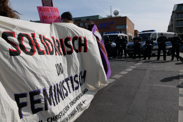 Eine Gruppe von Menschen marschiert auf einer Straße, hält eine Fahne mit der Aufschrift "Solidarität und Feminismus" hoch, mit parkenden Fahrzeugen und Gebäuden im Hintergrund unter einem klaren blauen Himmel.