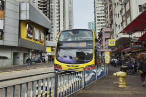 Ein gelber Doppeldeckerbus fährt eine Straße mit hohen Gebäuden entlang, mit Fußgängern auf der rechten Seite, einem Hydranten und einem Geländer, unter einem klaren blauen Himmel.