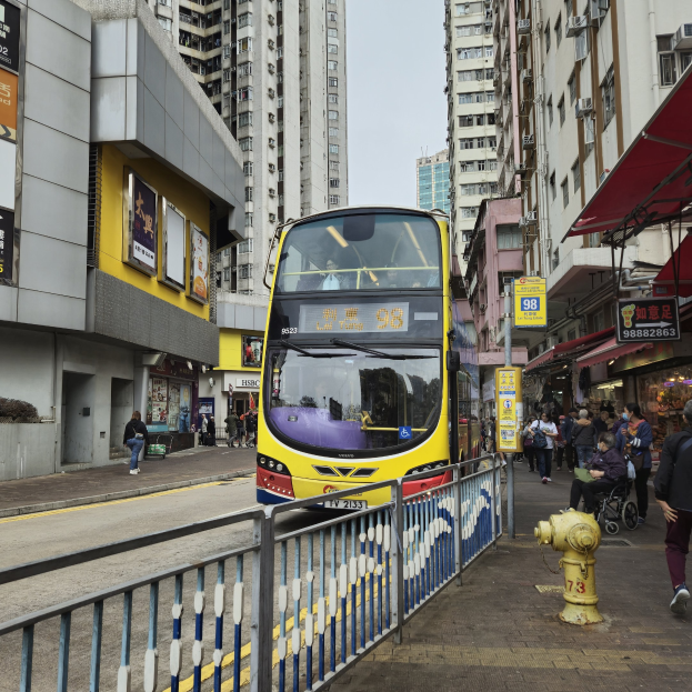 Ein gelber Doppeldeckerbus fährt eine Straße mit hohen Gebäuden entlang, mit Fußgängern auf der rechten Seite, einem Hydranten und einem Geländer, unter einem klaren blauen Himmel.