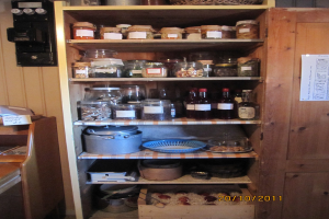 Eine Küchenszene mit Gläsern, Weinflaschen, Tellern, Platten, Körben und anderen Gegenständen auf Gestellen; ein Plakat an einer Tür rechts; ein elektronisches Gerät an der Wand oben links; und ein Tisch mit Papieren unten links.