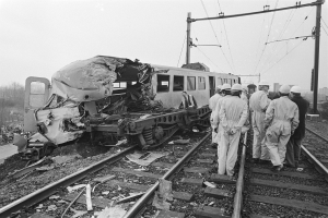 Eine Schwarz-Weiß-Szene eines Zugunglücks mit Menschen in Helmen, die herumstehen, Strommasten, Bäumen, Gebäuden und einem klaren Himmel im Hintergrund.