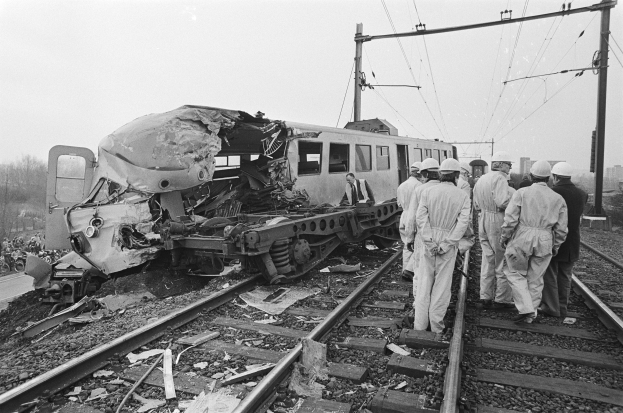 Eine Schwarz-Weiß-Szene eines Zugunglücks mit Menschen in Helmen, die herumstehen, Strommasten, Bäumen, Gebäuden und einem klaren Himmel im Hintergrund.