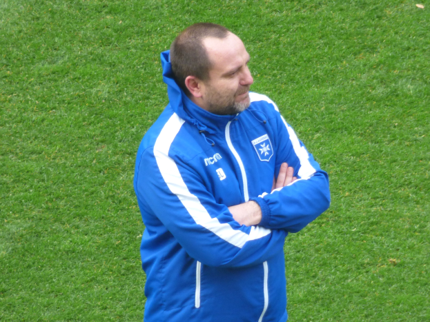 Ein Mann in einer blauen Jacke steht selbstbewusst auf einem grünen Feld und wirkt wie ein Trainer.