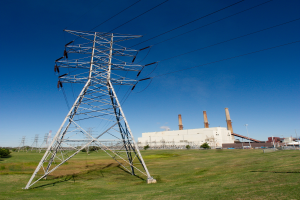 Strommasten und Kabel im Vordergrund mit Gebäuden, Schornsteinen, Bäumen und einem bewölkten Himmel im Hintergrund.