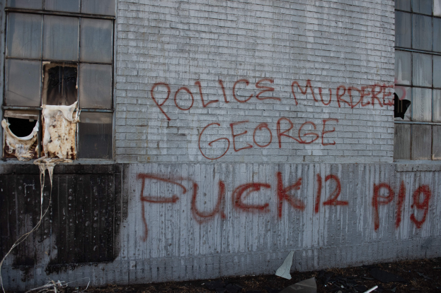 Ein Gebäude mit Graffiti, das 'Polizei Mord George Fuck 12 P19' auf der Seite trägt, mit Fenstern und verschiedenen Gegenständen vorne.