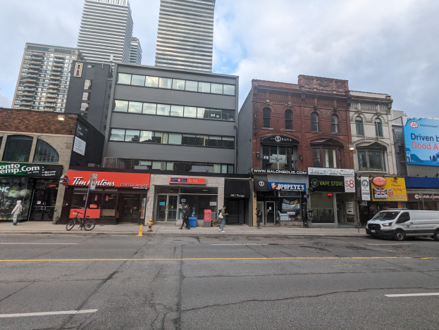 Stadtstraße mit hohen Gebäuden, fahrenden Fahrzeugen, Fußgängern, einem Fahrrad, einem Hydranten und Werbetafeln für Mietwohnungen unter einem bewölkten Himmel.