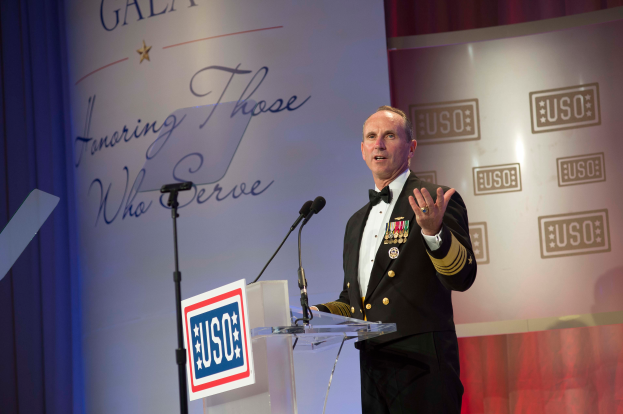 Ein Mann in militärischer Uniform steht an einem Podium mit zwei Mikrofonen und einer Tafel und hält eine Rede hinter einem Banner und einem Vorhang.