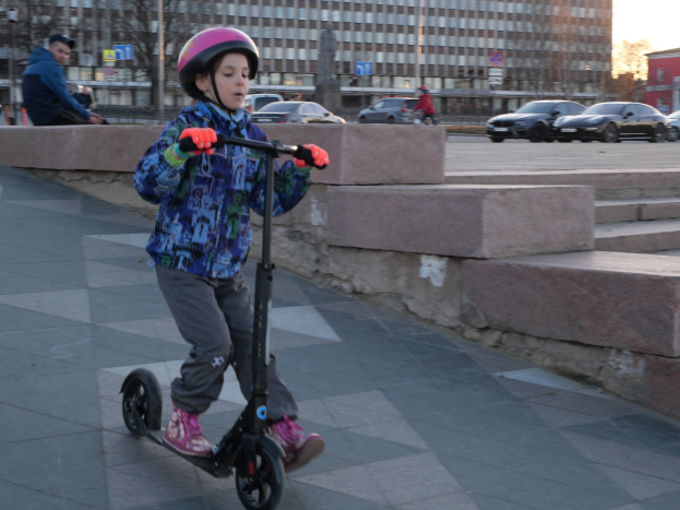 Ein junger Junge mit Helm und Handschuhen fährt auf einem Gehweg mit Stufen, Fahrzeugen, Menschen, Bäumen, Pfählen, Brettern, Gebäuden und einem klaren blauen Himmel im Hintergrund.