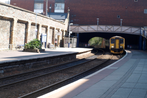 Ein Zug fährt auf den Schienen mit Plattformen auf beiden Seiten, eine Hauswand links, Straßenlaternen, Pflanzen, Bänke und eine Kabine vor der Wand, eine Brücke über die Plattformen und Gebäude und Masten im Hintergrund.