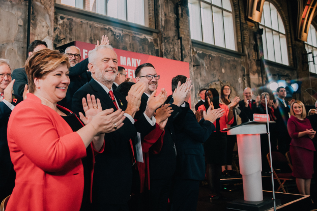 Eine Gruppe von Menschen, die vor einem Publikum stehen und jubeln, mit einem Podium, einem Mikrofon und einer Tafel mit Text auf der rechten Seite und Stühlen, einer Fahne, einer Wand, Fenstern und Lichtern im Hintergrund.