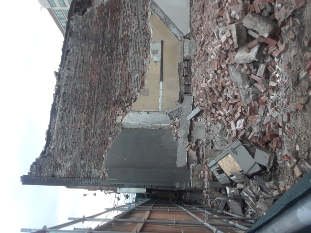 Ein teilweise abgerissener Bau mit verstreuten Ziegeln und Metallstangen, umgeben von anderen Gebäuden unter einem klaren blauen Himmel.