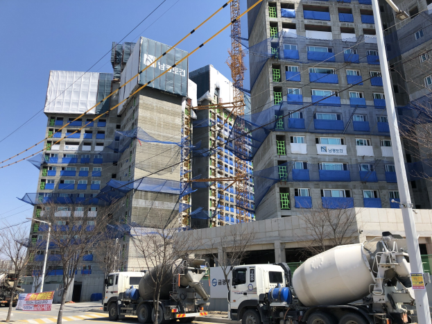 Baustelle mit einem Betonmischer-Lkw, der vor einem hohen Gebäude im Bau geparkt ist, Fahrzeugen auf der Straße, Bäumen, Strommasten, Brettern, Kabeln im Vordergrund und Himmel im Hintergrund.