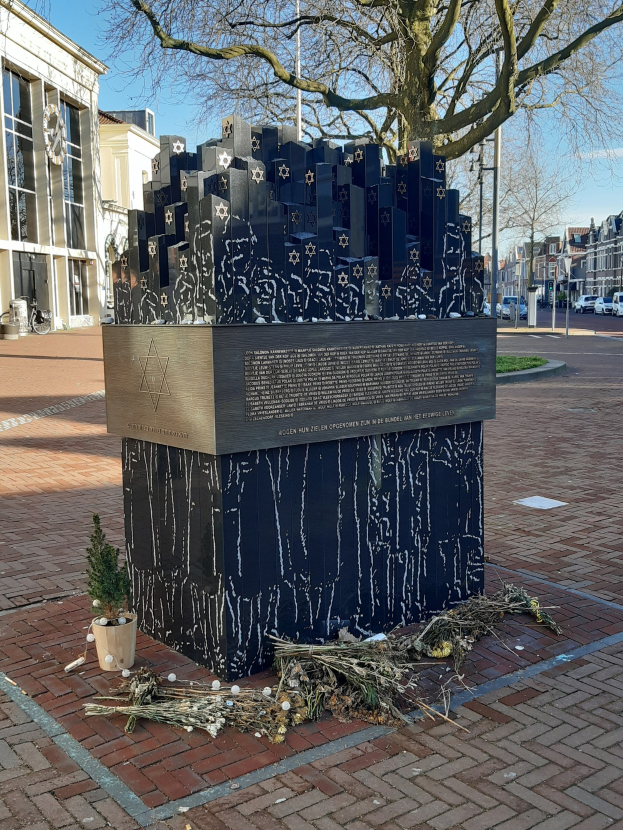 Denkmal für Holocaust-Opfer in einem Stadtplatz mit einem Baum davor, einer Topfpflanze, verstreuten trockenen Blättern und Gebäuden im Hintergrund.