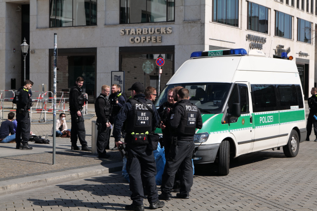 Eine Gruppe von Polizeibeamten steht vor einem Starbucks-Café mit einem Van auf der rechten Seite und einigen Menschen auf der linken Seite, während im Hintergrund ein Gebäude, ein Schild, ein Laternenmast und ein Zaun zu sehen sind.