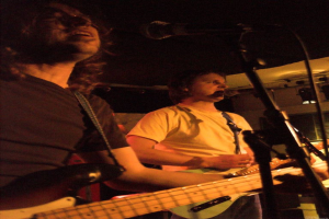 Eine Person in einem weißen Hemd singt in ein Mikrofon und spielt eine grüne Gitarre, steht neben einer anderen Person in einem schwarzen Hemd und Brille, die ebenfalls singt und eine schwarze Gitarre spielt, mit Scheinwerfern und anderen Instrumenten im Hintergrund.