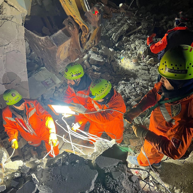 Gruppe von Menschen in orangen Uniformen und grünen Helmen bei der Arbeit an Schutt mit einem Kran im Hintergrund bei einem Einsatz nach dem Einsturz eines Gebäudes.