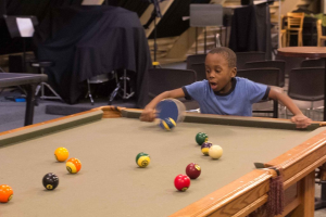 Ein Junge steht mit einem Tischtennis-Schläger vor einem Snookertisch mit Billardkugeln, im Hintergrund sind Stühle und andere Gegenstände zu sehen.
