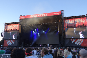 Eine große Menschenmenge steht vor einer hell erleuchteten Bühne beim Shaky Knees Music Festival, wo Musiker unter Bannern spielen, mit einem Geländer im Vordergrund und einem klaren blauen Himmel im Hintergrund.