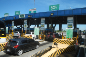 Eine Mautstation mit parkenden Autos davor, Fahrzeugen auf der Straße, Verkehrskegeln, Schildern mit Text, einer Brücke, Pfählen, Drähten, Bäumen und einem klaren blauen Himmel im Hintergrund.