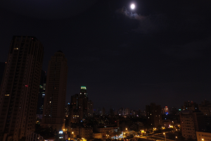 Eine nächtliche Stadtansicht mit hohen Gebäuden, Häusern und zahlreichen Straßenlaternen, mit einem mondhellen Himmel im Hintergrund.