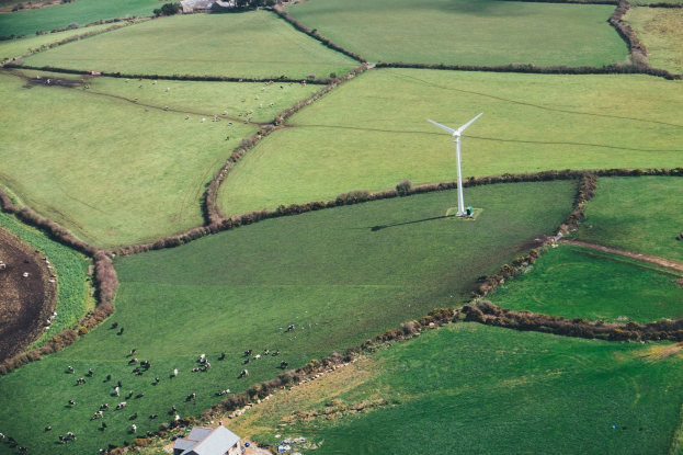 Luftaufnahme einer Windkraftanlage in einer grünen Wiese mit Bäumen, Häusern und Tieren in Irland.
