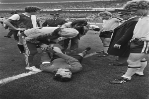 Schwarzes und weißes Bild eines Fußballspielers, der auf dem Boden liegt und von einer Gruppe Männer umgeben ist, mit einer Menge im Hintergrund eines Stadions.