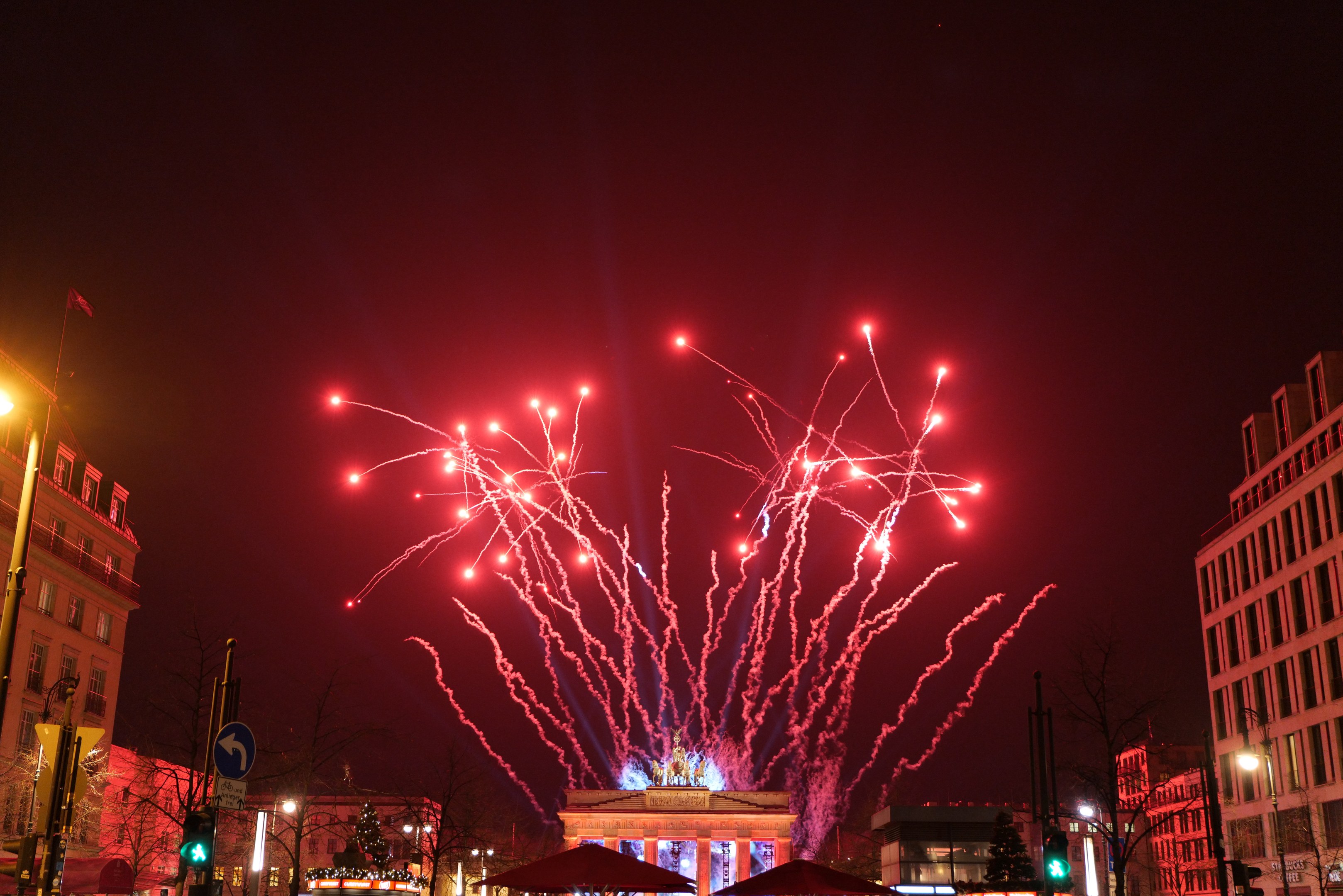 Eine belebte Stadtstraße an einem Silvesterabend in Berlin mit Gebäuden, Bäumen, Laternenmasten, Ampeln, Schildern, Zelten, Menschen und einem prächtigen Feuerwerk am Himmel.