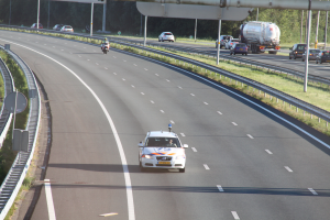 Ein Auto fährt auf einer Autobahn mit einem polizeilichen Fahrzeug am Rande, anderen Fahrzeugen, Leitplanken, Pfölen, Planken und Bäumen im Hintergrund.