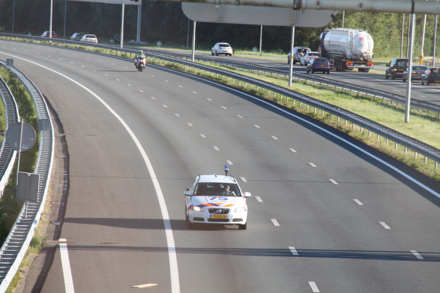 Ein Auto fährt auf einer Autobahn mit einem polizeilichen Fahrzeug am Rande, anderen Fahrzeugen, Leitplanken, Pfölen, Planken und Bäumen im Hintergrund.