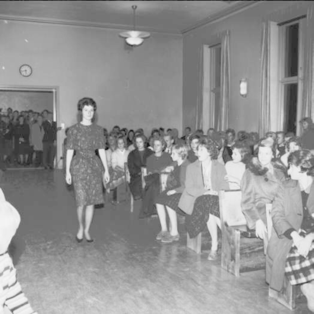 Eine Frau in einem schwarzen und weißen Kleid läuft über einen Laufsteg während einer Modenschau für Frauenwahlrecht, mit einer Menge, die auf Stühlen sitzt und einer Uhr und Lampen an der umliegenden Wand.