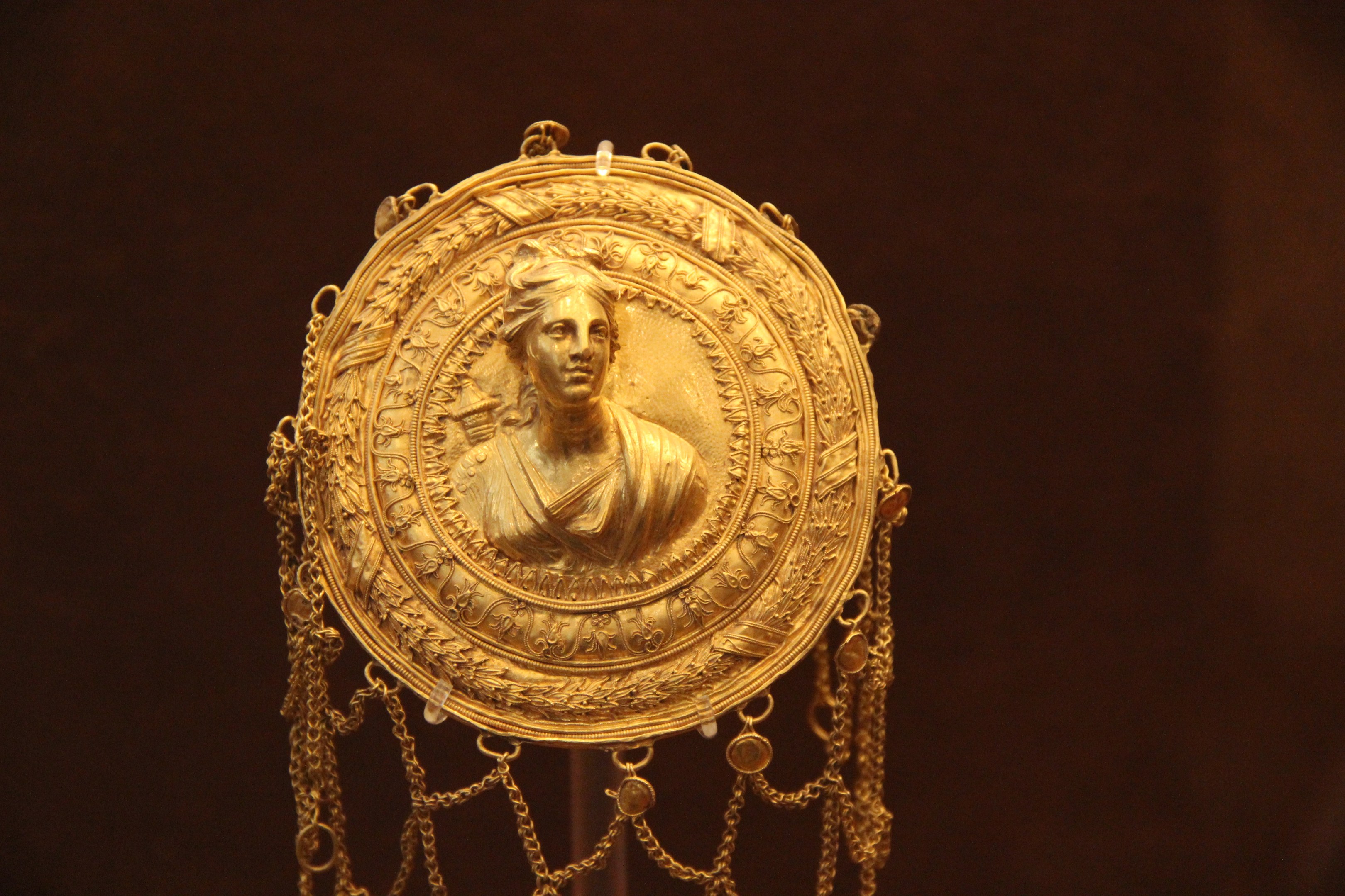 Goldene Medaille mit einer Frauengestalt, die detailliert verziert ist und vor einem dunklen Hintergrund zu sehen ist.