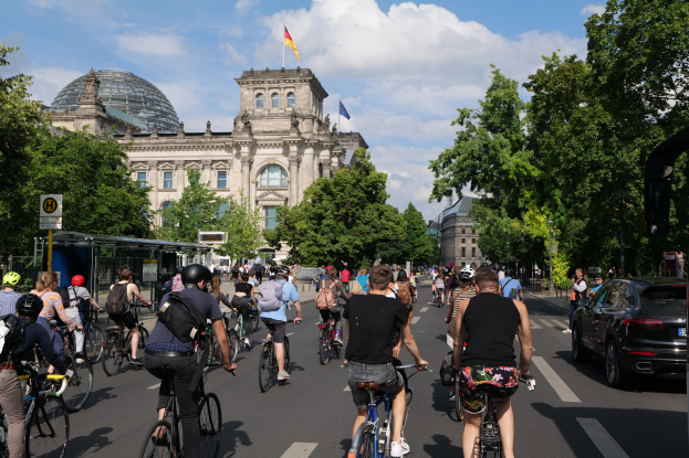 Gruppe von Menschen, die auf Fahrrädern eine von Bäumen gesäumte Straße in Berlin, Deutschland, entlangfahren, mit Gebäuden und einer Bushaltestelle, unter einem bewölkten Himmel und einer Flagge, die auf einem Gebäude weht.