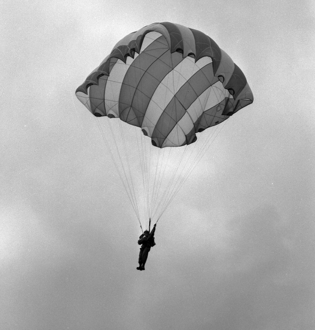 Eine Person beim Drachenfliegen am Himmel mit einem Fallschirm vor einem bewölkten Hintergrund, in Schwarzweiß aufgenommen.