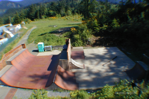 Eine Skaterbahn mit Kisten auf der linken Seite, Bäumen, Gras, Pflanzen und Blumen im Hintergrund und Text unten rechts.