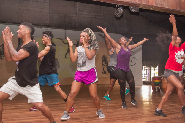 Eine Gruppe von Menschen, die in einem Studio mit einer bemalten Wand, einer Glasscheibe und Deckenbeleuchtung tanzen, wahrscheinlich während eines Zumba-Kurses.