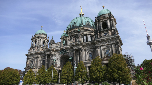 Berliner Dom, eine große Kathedrale mit Fenstern, Säulen, Bögen und Statuen, vor einem bewölkten Himmel mit Menschen, Bäumen und einem Turm im Vordergrund.