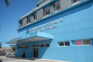 Krankenhaus- und Medizinzentrum Carmona mit Text darauf, umgeben von Bäumen, Fahrzeugen und anderen Gegenständen, unter einem klaren blauen Himmel.