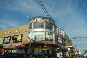 Eine belebte Stadtstraße mit fahrenden Autos, Fußgängern auf dem Gehweg und einem großen Gebäude mit der Aufschrift "Royal Mall" im Hintergrund, umgeben von parkenden Autos, Bäumen, Strommasten und Drähten unter einem Himmel.