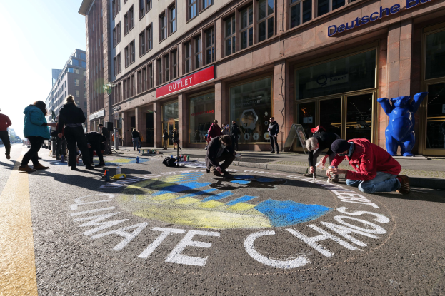 Eine Gruppe von Menschen sitzt vor einem Gebäude mit Fenstern und Namensschildern auf dem Boden, umgeben von Flaschen und anderen Gegenständen, mit Bäumen und einem klaren blauen Himmel im Hintergrund.