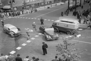 Eine Schwarz-Weiß-Szene eines Autounfalls an einer Straßenecke mit mehreren Fahrzeugen, darunter einem Van, einer Gruppe von Menschen drumherum und im Hintergrund Gebäude, Bäume, Masten und Texttafeln.