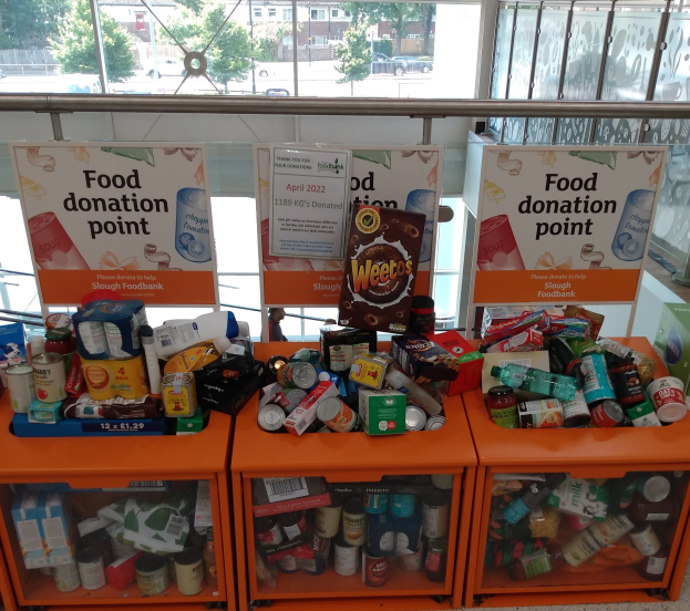 Orange Schränke voller Lebensmittelspenden, darunter Flaschen, Dosen und Kartons, mit Informationsschildern und Blick auf Fahrzeuge, Bäume, Gebäude und Himmel durch Glasfenster.