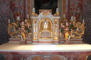 Ein Altar in einer Kirche mit Skulpturen von Menschen und Kerzen auf St"nden, vor einer Wand mit einem Bild.