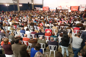 Ein volles Stadion mit vielen sitzenden und stehenden Menschen, eine Bühne mit zwei Rednern an einem Podium und zahlreiche Banner und Lichter.