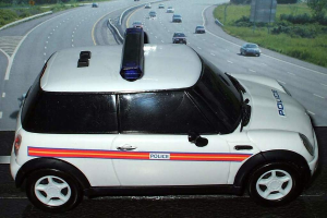 Ein weißer Polizeiwagen ist im Vordergrund geparkt, mit mehreren Autos, die auf der Straße dahinter fahren, umgeben von Gras und Bäumen auf beiden Seiten in einer ländlichen Umgebung.