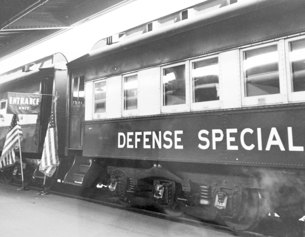 Schwarzes und weißes Foto eines Zuges mit der Aufschrift "Defense Special" auf den Eisenbahnschienen mit zwei Fahnen links und einer Decke oben.